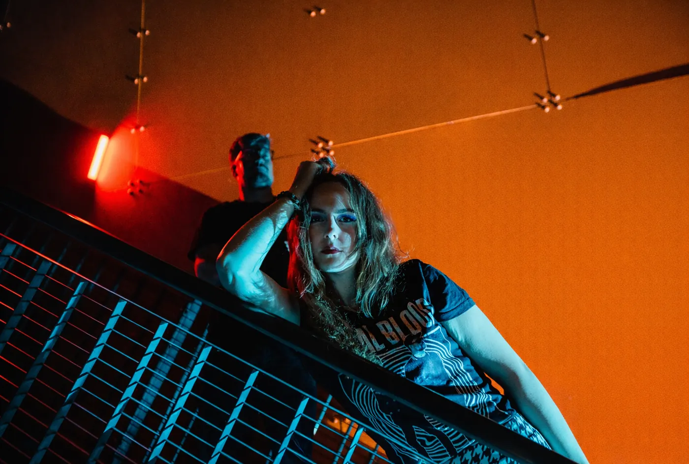 Ronja Kaminsky leaning on a railing in front of an orange wall, Nils Degenhardt behind her. Photo: Aloha Burn